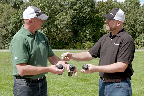 Two adult males wearing polo shirts and billed caps stand outdoors, talking to each other as they hold irrigation rotors in their hands. Grass and shrubbery are in the background.