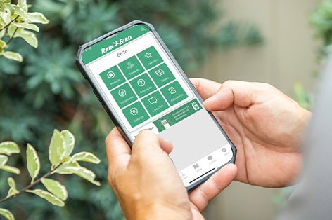 A pair of adult hands hold a smartphone that displays the Rain Bird Resources app on its screen. Shrubbery is in the background.