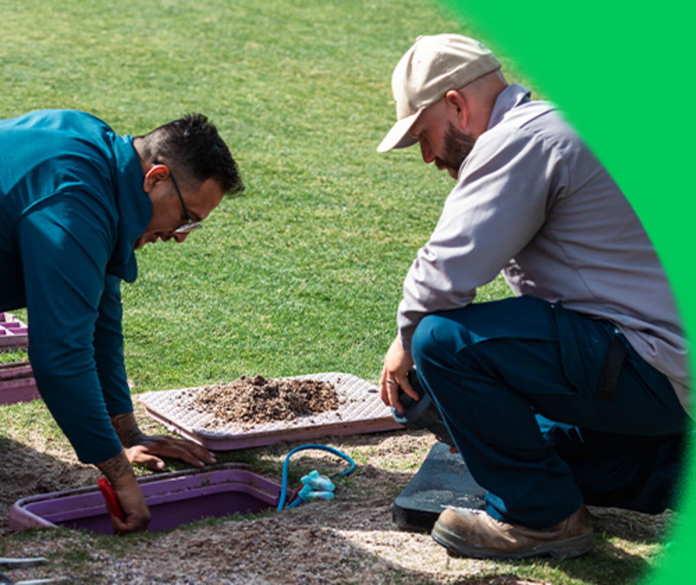 Two adult male irrigation professionals kneel outdoors on grass as they examine an in-ground irrigation access panel that has been opened.